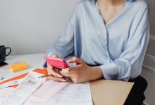 Mulher usa uma calculadora rosa entre contas e recibos sobre uma mesa.