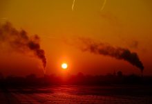 Chaminés de um complexo industrial emitindo fumaça sobre a paisagem ao entardecer.