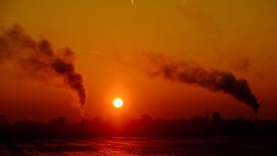 Chaminés de um complexo industrial emitindo fumaça sobre a paisagem ao entardecer.