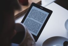 Pessoa lê em um leitor digital Kindle enquanto segura uma xícara de café, em ambiente interno.