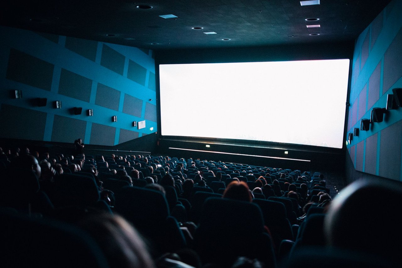 Público lotado sentado em uma sala de cinema enquanto assiste a um filme exibido em uma tela grande.