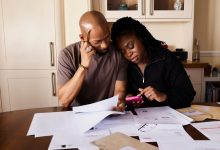 Casal analisa documentos e usa um celular enquanto administra as finanças domésticas à mesa.
